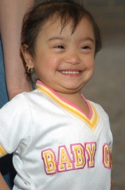 A toddler girl with Down Syndrome smiles proudly for the camera
