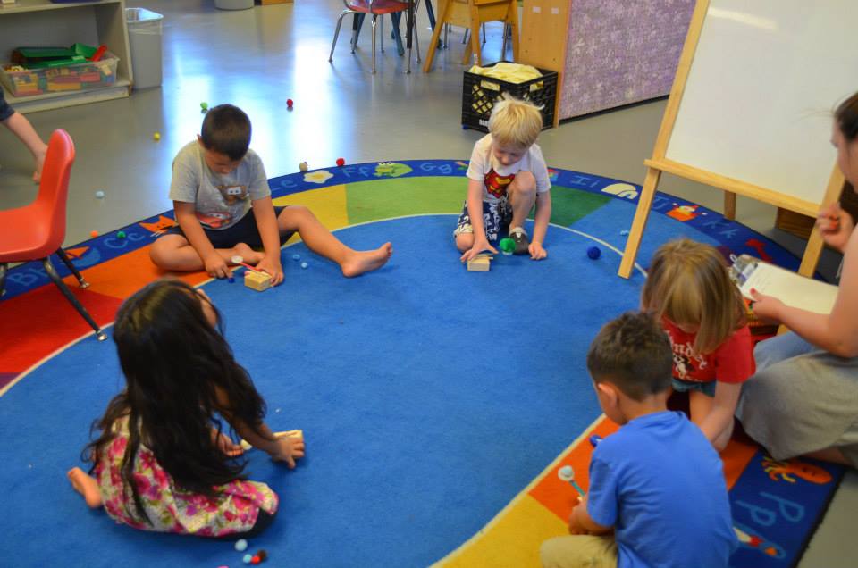 Children at the Child Development Center launch pom-poms from their catapults across a blue oval rug