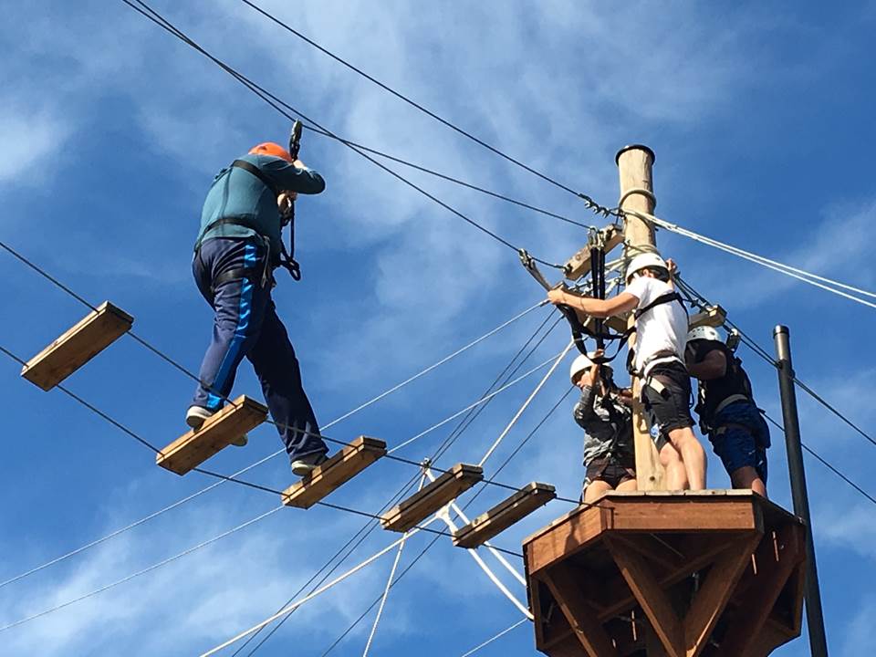 Four students at a ropes course. Three students are standing on a small platform high in the air, while the fourth student crosses to them on a ladder bridge.